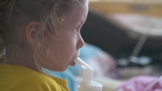 pretty small girl in yellow t-shirt breathes with inhaler lying in bed in light room at home extreme close view