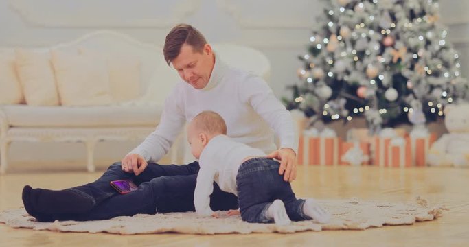 Pretty baby crawls on the carpet to dad and sits on his knees. Cheerful dad is watching cartoons with his little son on the phone. Merry winter holidays at the Christmas tree.
