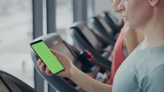 Athletic Woman Does Gestures while Using Green Screen Smartphone and Running on the Treadmill in a Gym. Fit Female Posts on Social Media, Takes Pictures, Watches Videos, Exercises in Fitness Club