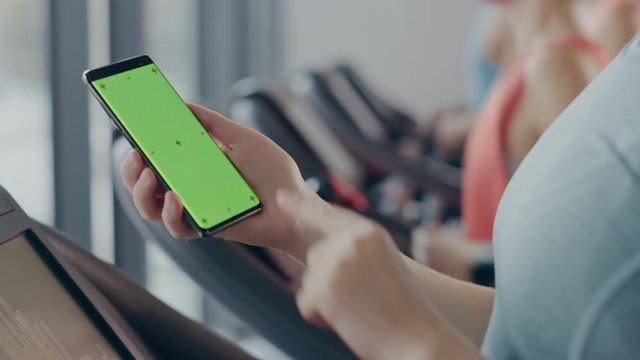 Athletic Woman Does Gestures while Using Green Screen Smartphone and Running on the Treadmill in a Gym. Fit Female Posts on Social Media, Takes Pictures, Watches Videos, Exercises in Fitness Club