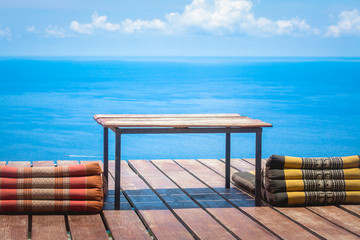 Table and Thai Triangle Pillows on Wooden Terrace with Tropical Beach, Blue Sky and Clouds in Background
