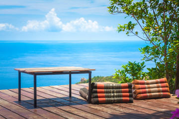 Table and Thai Triangle Pillows on Wooden Terrace with Tropical Beach, Blue Sky and Clouds in Background
