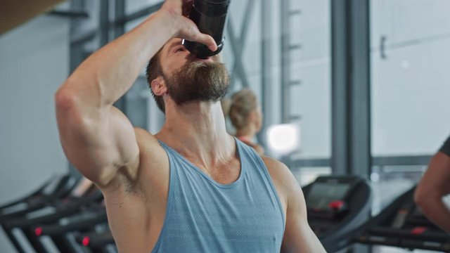 Muscular Heavyweight Champion Walks Through Gym, Drinks Protein Cocktail from Tumbler for Hydration and Muscle Mass. In the Background Sports People Running on Treadmills