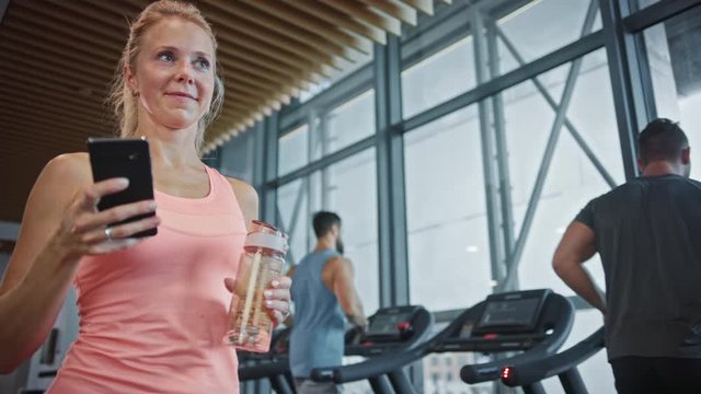 Beautiful Athletic Woman Uses Smartphone Walks Through Fitness Club Gym. Using Social Media, Posting Pictures, Communicating, Checking Email. In the Background Sports People Running on Treadmills