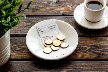 Pay restaurant bill by cash. Reciept and coins on plate on dark wooden background