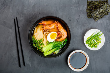 Asian noodle soup, ramen with prawn shrimp, vegetables and egg in black bowl on gray concrete...