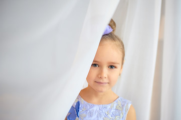 Portrait of a little cute smiling girl peeking out from behind a white curtain.
