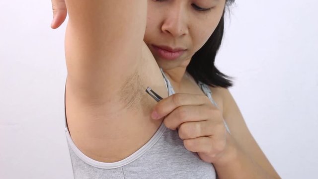 Close up of young woman plucking armpit underarm with tweezers isolated on grey background. Concept of Hygiene skin body care and beauty.
