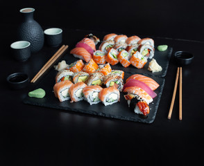Various kinds of classic sushi and rolls served on black stone with bottle of sake on black background.