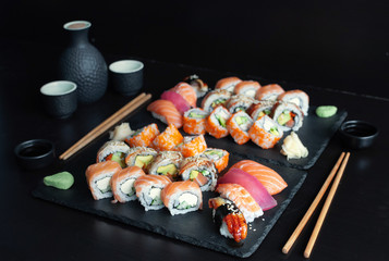 Various kinds of classic sushi and rolls served on black stone with bottle of sake on black background.