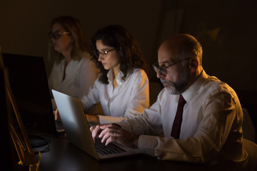 Fototapeta na wymiar Serious businessman and businesswomen using computers. Side view of professional male and female business colleagues using laptop computers in dark office. Working late concept