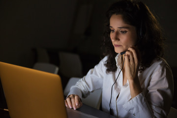 Serious woman with headset using laptop. Focused female call center worker working with laptop late...