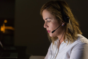 Female call center operator in dark office. Side view of concentrated businesswoman in headset...