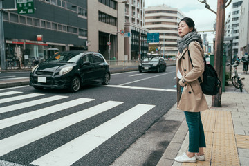 pretty asian chinese woman traveler walking in city osaka waiting for traffic lights to cross busy road with cars driving by. full length girl with bag standing on street and ready to pass pedestrian