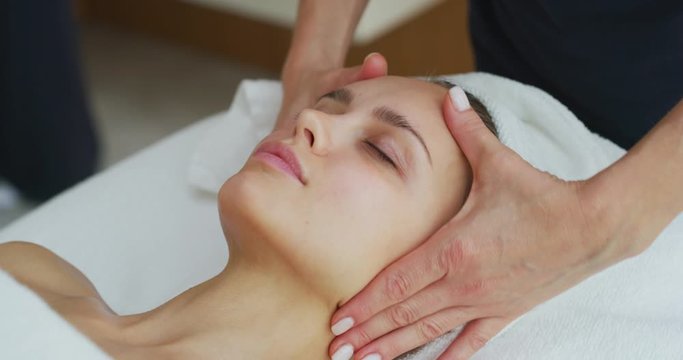 Close up of a beautiful young smiling woman with a towel on her head is receiving a facial massage and spa treatment for perfect skin in a luxury wellness center.