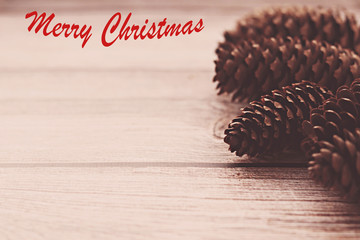 Brown pine cones on a wooden background with the inscription Merry Christmas in red. The bumps look like a silhouette. Chocolate toning. Selective focus. Decorative Christmas background.