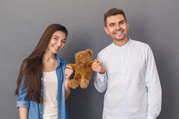 Relationship Concept. Young couple studio standing isolated on grey with teddy bear laughing joyful