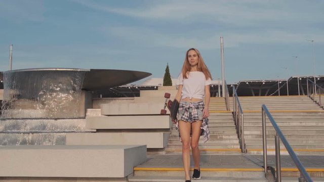 A young blonde girl, a model with a longboard in her hand in a white shirt and denim shorts goes down the stairs and poses for the camera. Urban modern structure in the background.