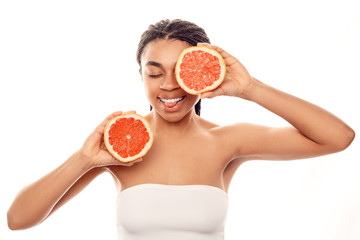 Beauty Concept. Young african woman wearing top isolated on white covering eye with grapefruit half laughing showing tongue cheerful