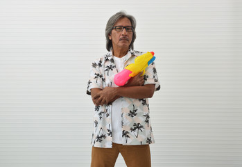 Senior traveler asian man wearing glasses, summer shirt and brown shorts holding colorful water gun over white wall background, Business summer holiday concept