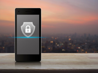 Padlock with shield flat icon on modern smart mobile phone screen on wooden table over blur of cityscape on warm light sundown, Business security insurance online concept