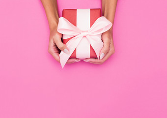 Woman hands holding present box with a ribbon on pastel pink background .