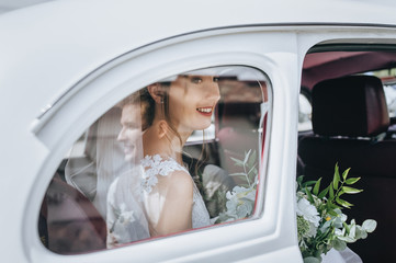 A beautiful bride with curly hair and a bouquet sits near a window in a white car, and the groom is reflected. Wedding portrait close-up newlyweds. Photography and concept.