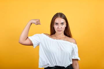 Strong charming young woman showing muscules, looking serious isolated on orange background in studio in casual white shirt. People sincere emotions, lifestyle concept.