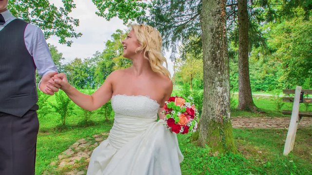 A dynamic front close shot of newlywed walking down a curve path holding hands surrounded by trees and grass smiling toward each other.