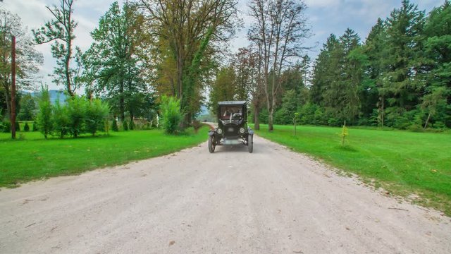 A slow-motion dynamic dolly shot of a black vintage car driving through a path surrounded by nature.