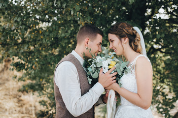 Wedding portrait close-up of beautiful newlyweds who enjoy a bouquet. Stylish groom in a suit gently looks at a cute bride in a white dress on a background of nature. Photography and concept.