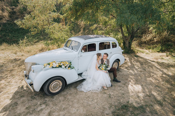 Obraz na płótnie Canvas Beautiful newlyweds are sitting and hugging near an old retro car and summer nature. Wedding portrait of a stylish, smiling groom and lovely bride with curly hair. Photography and concept.