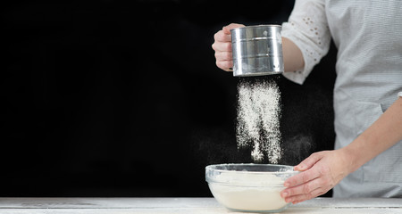 Woman sifts flour through a sieve in a glass bowl. Empty space for text. Isolated on dark background