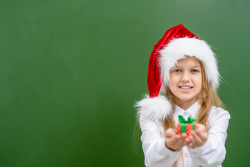 Smiling girl wearing a red christmas hat holds gift box near green chalkboard. Empty space for text