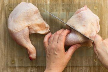 Woman hands cutting raw chicken leg in two part with knife raw on glass cutting board on wooden...