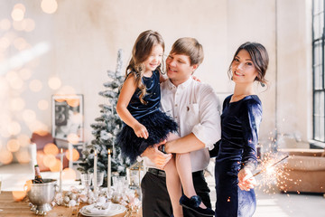 Happy family in blue - woman, man and little girl, with a sparkler in hand, near the New Year's table for Christmas at home. The girl at the father on hands. New year 2020.