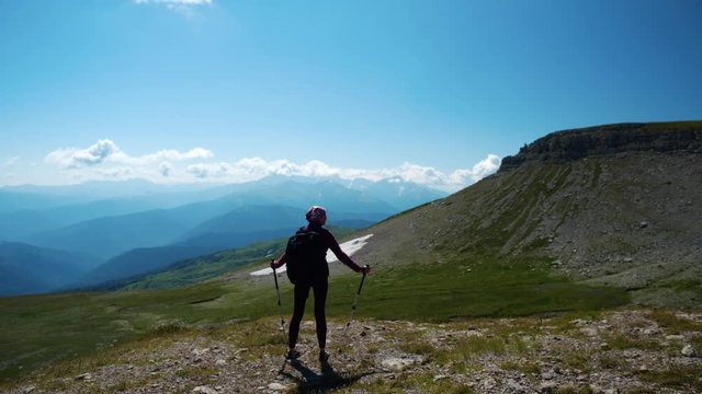 Lady hikes in beautiful Caucasian mountains. Young woman with backpack and trekking poles on the way to the mountain top makes a pause to observe fantastic view from a height.