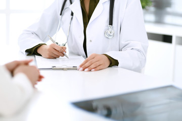 Woman doctor and patient sitting and talking at medical examination at hospital office, close-up. Therapist filling up medication history records. Medicine and healthcare concept