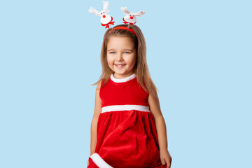 Little Girl on Blue Background. Isolated. Happy emotional Child in a Red Dress, a Girl wears a Christmas Hoop with Santa's deer on her Head. Child waving a Red dress. little girl with party hat  