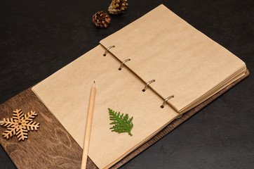 Notebook in wooden cover with kraft blank sheets, pencil, fir tree branch and wooden snowflake on black background. Plan next year concept. To do list. Christmas background. Mockup. Copy space