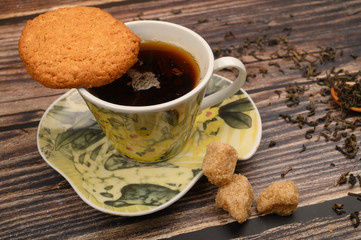 A Cup of black tea, tea leaves, pieces of brown sugar, oatmeal cookies on a wooden background. Close up.