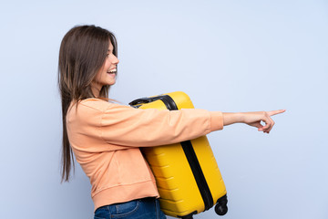 Traveler Brazilian girl holding a suitcase over isolated blue background pointing to the side to present a product