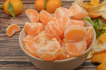 Peeled tangerines in a ceramic dish, tangerines on a twig with green leaves, tangerine peel on a wooden background. Autumn harvest. Close up.