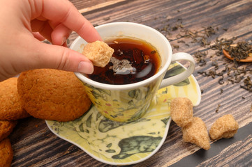 The girl's hand puts a piece of brown sugar in a mug of black tea, oatmeal cookies, tea leaves, brown sugar on a wooden background. Close up.