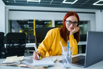 Young girl works at a computer in the office. Beautiful freelancer working from a laptop computer. Teenager student at the computer listens music to earphones and prepares for exams. Business woman.