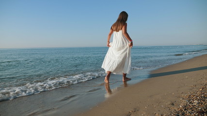 A young girl in a white dress barefoot walks along the sea coast in the morning, slow-motion rear view shot