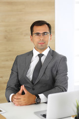 Cheerful smiling businessman sitting at a desk in modern office. Headshot of male entrepreneur or company director at workplace. Business concept