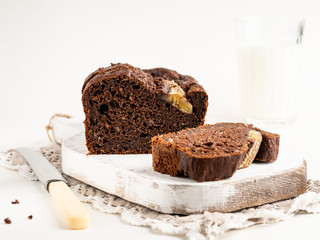 Gorgeous tasty chocolate banana cake (banana bread) sliced and decorated with banana split on white wooden cutting board with a glass of milk, white background
