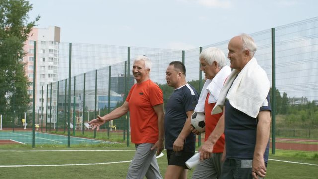 Side view of group of five tired multiethnic elderly men having conversation with each other while walking away from football field after game