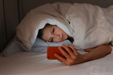 Young woman can't sleep and using her smartphone in beg at night. Insomia and unhealthy sleep because of gadget and Internet dependence. Social networks addiction concept